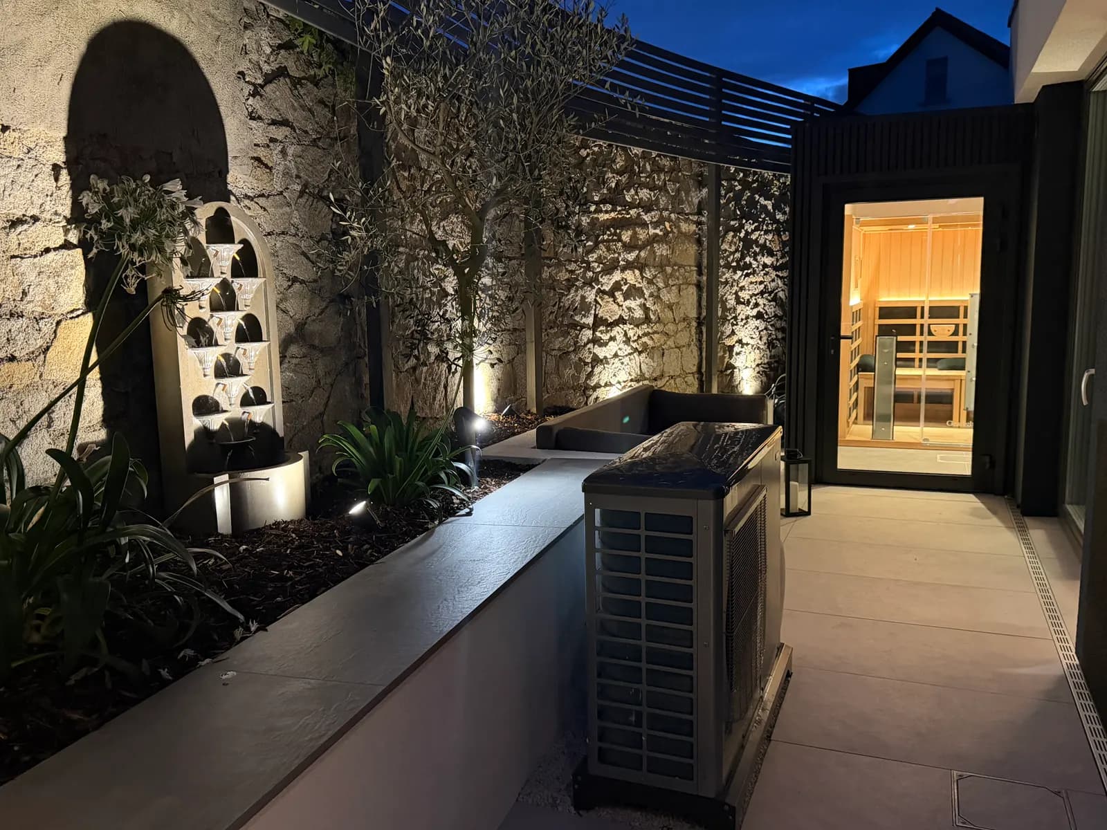 Garden wellness area at night with outdoor lighting, planting and glowing sauna